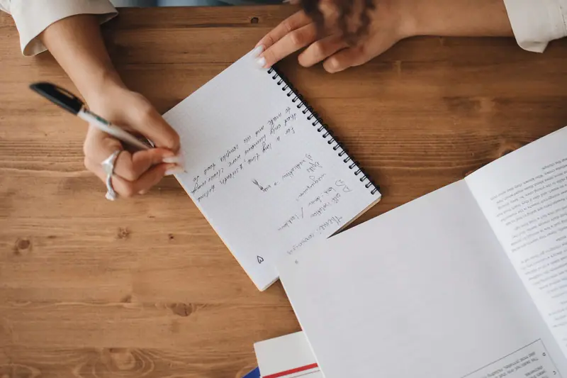 Hånd som skriver notater i en notatbok ved siden av en åpen bok